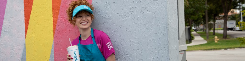A cafe employee enjoying a Tropical Smoothie Cafe Smoothie dressed in a pink tshirt, a blue cap & a blue apron branded with Tropical Smoothie Cafe Logo. "Apply to your local cafe"